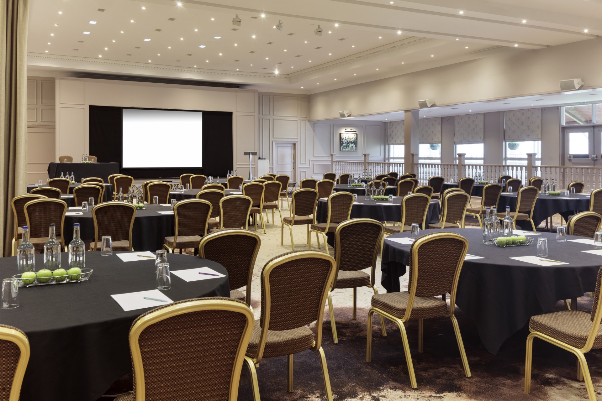 A large conference hall set up cabaret-style with round tables covered in black linens, gold-framed chairs, notepad settings with water bottles, and a large projection screen on a raised stage at the front.