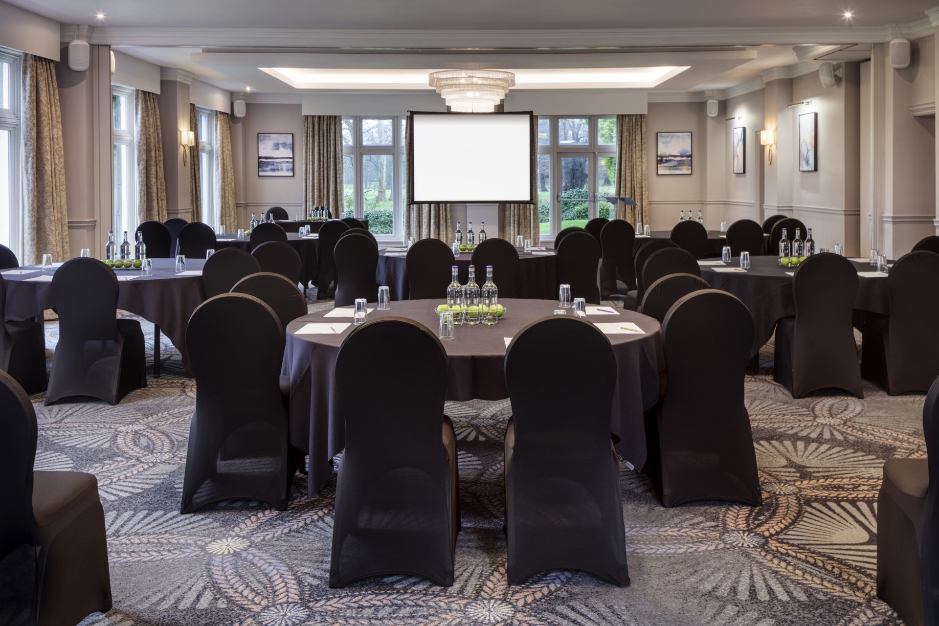 A conference and event room set up in cabaret style, featuring round tables with black tablecloths and matching black chair covers, equipped with glass water bottles, green apples, and a large central projector screen.