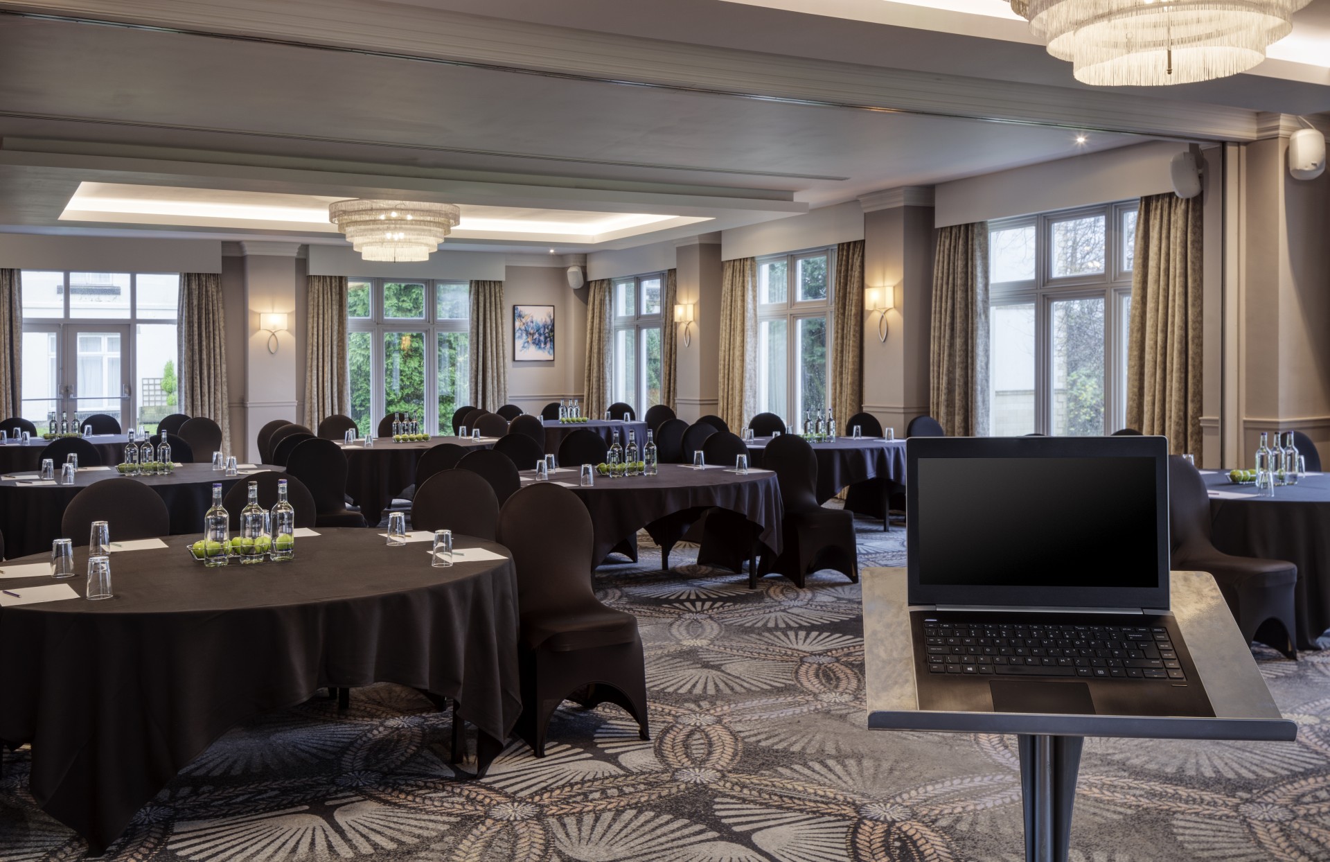 A modern conference room prepared for a presentation, featuring round tables with black tablecloths and black chair covers, crystal chandeliers, large windows, and a podium in the foreground with an open laptop.