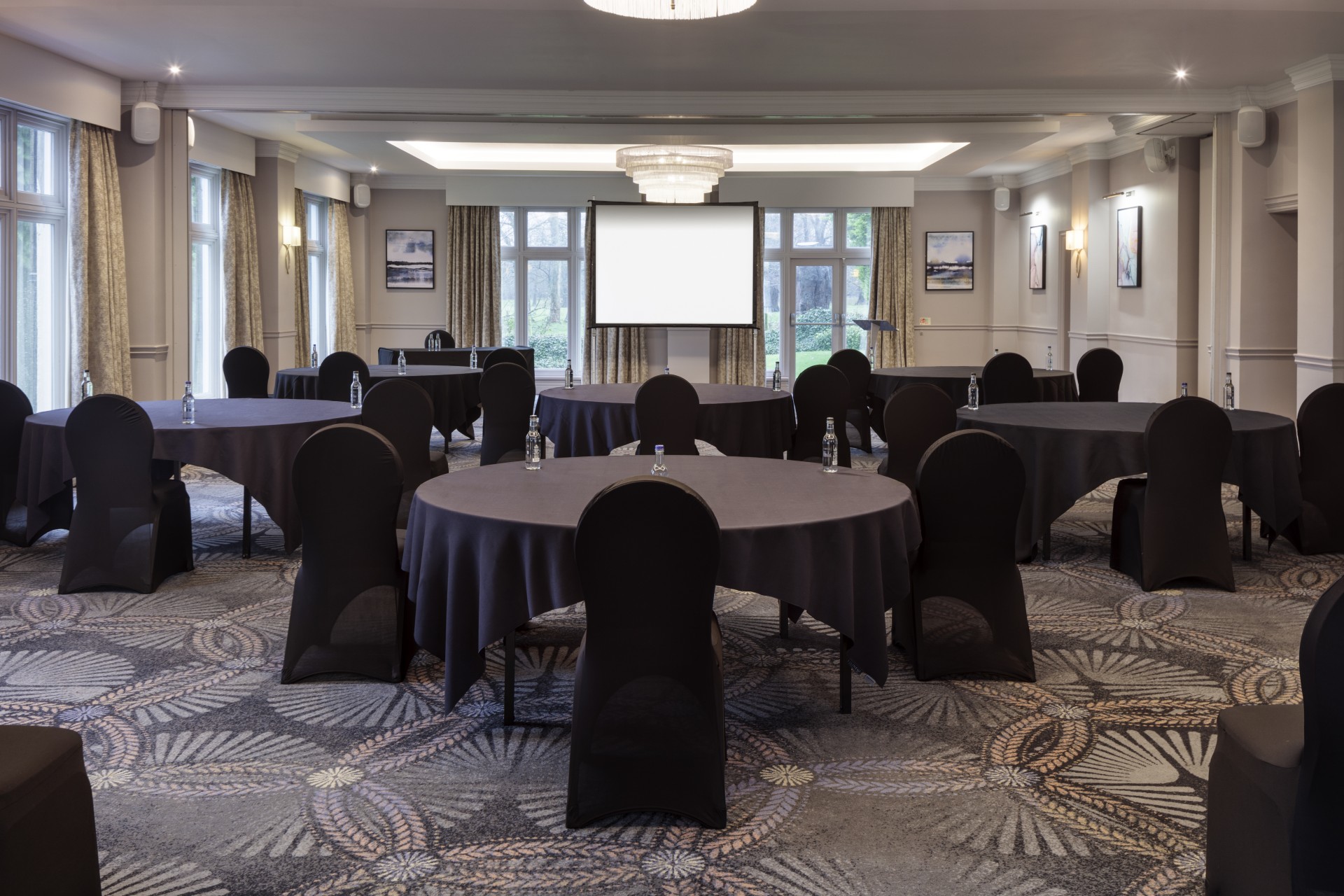 A carpeted meeting room arranged with round tables in dark tablecloths, black chair covers, water bottles, chandelier lighting, and a portable projection screen at the far end.
