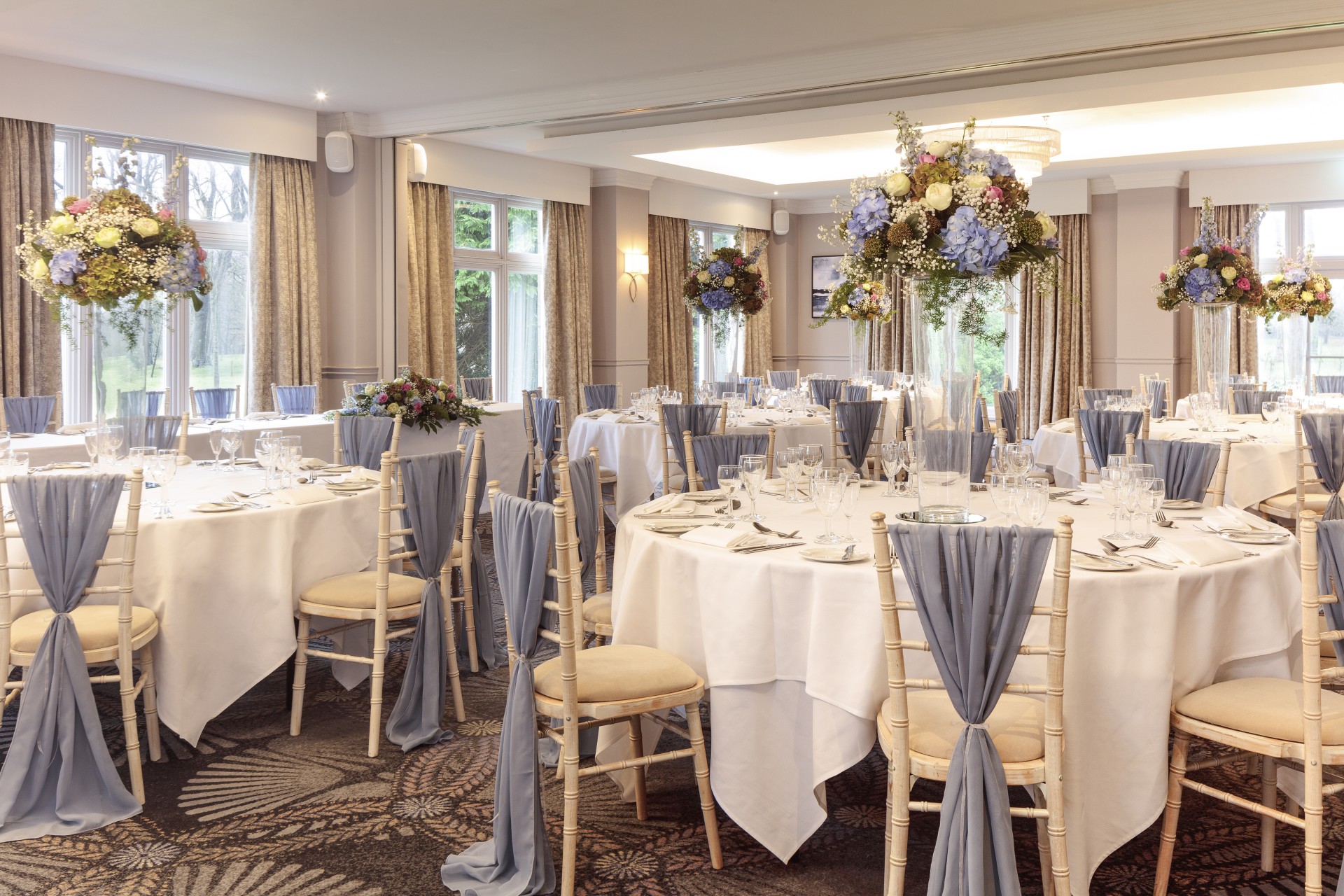 A bright banquet hall decorated for a wedding reception, featuring round tables with white linens, tall floral centerpieces with blue hydrangeas, and light wooden Chiavari chairs adorned with dusty blue fabric sashes.