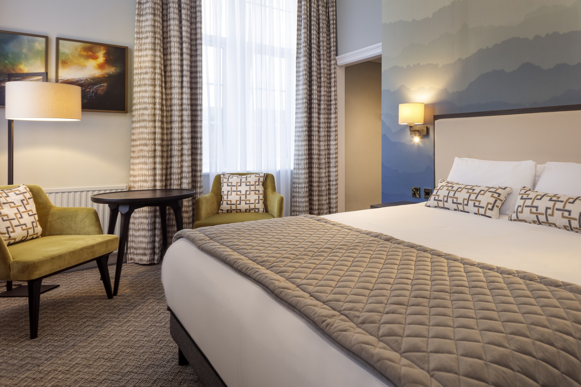 A stylish hotel bedroom featuring a bed dressed with a beige quilted runner and patterned pillows, two mustard-green armchairs with a small coffee table near a curtained window, and a blue mountain-mural accent wall.