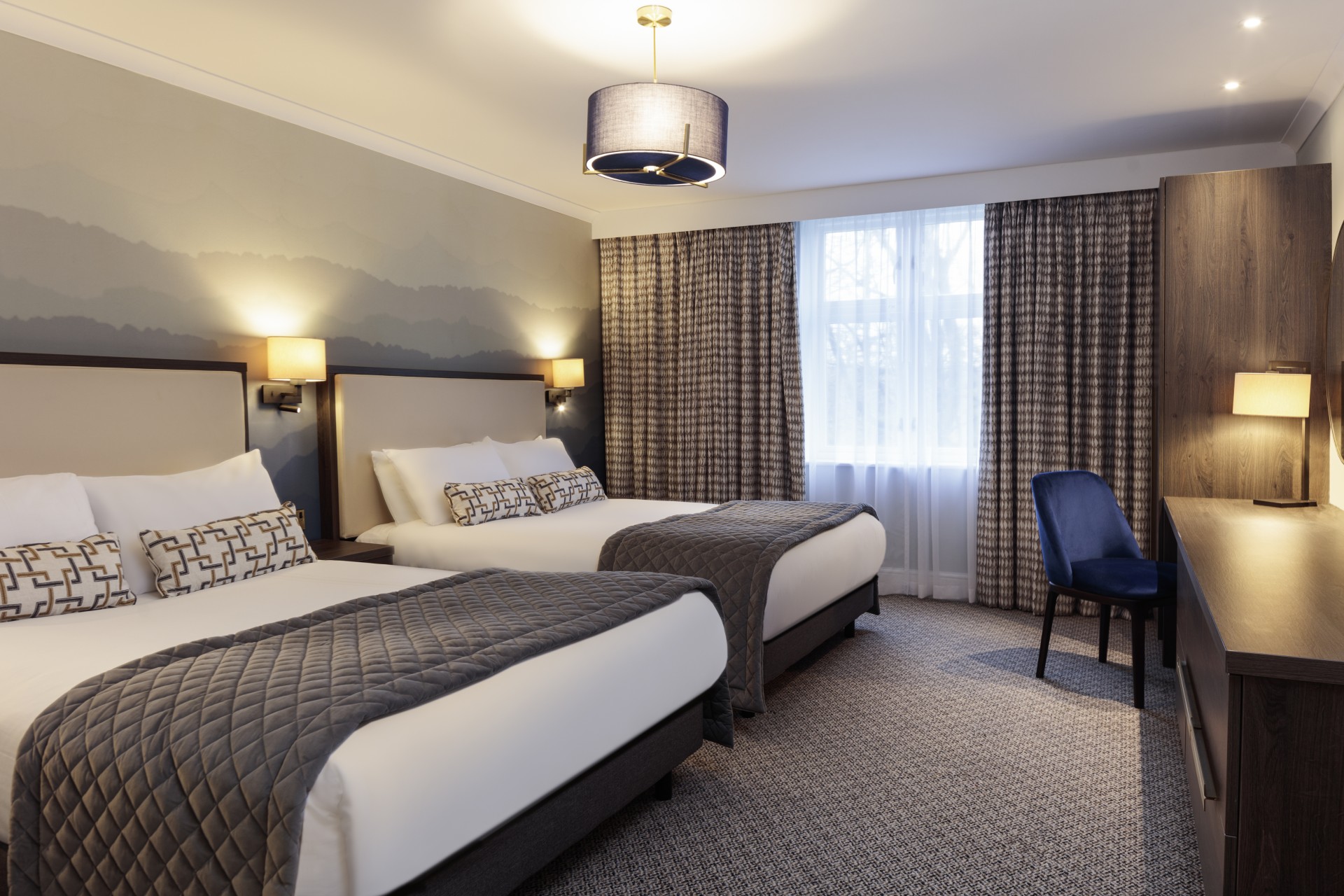 A modern hotel bedroom with two double beds dressed in white linens and grey quilted runners, a mountain-mural accent wall, dark wooden furniture, and a blue upholstered chair at a desk.
