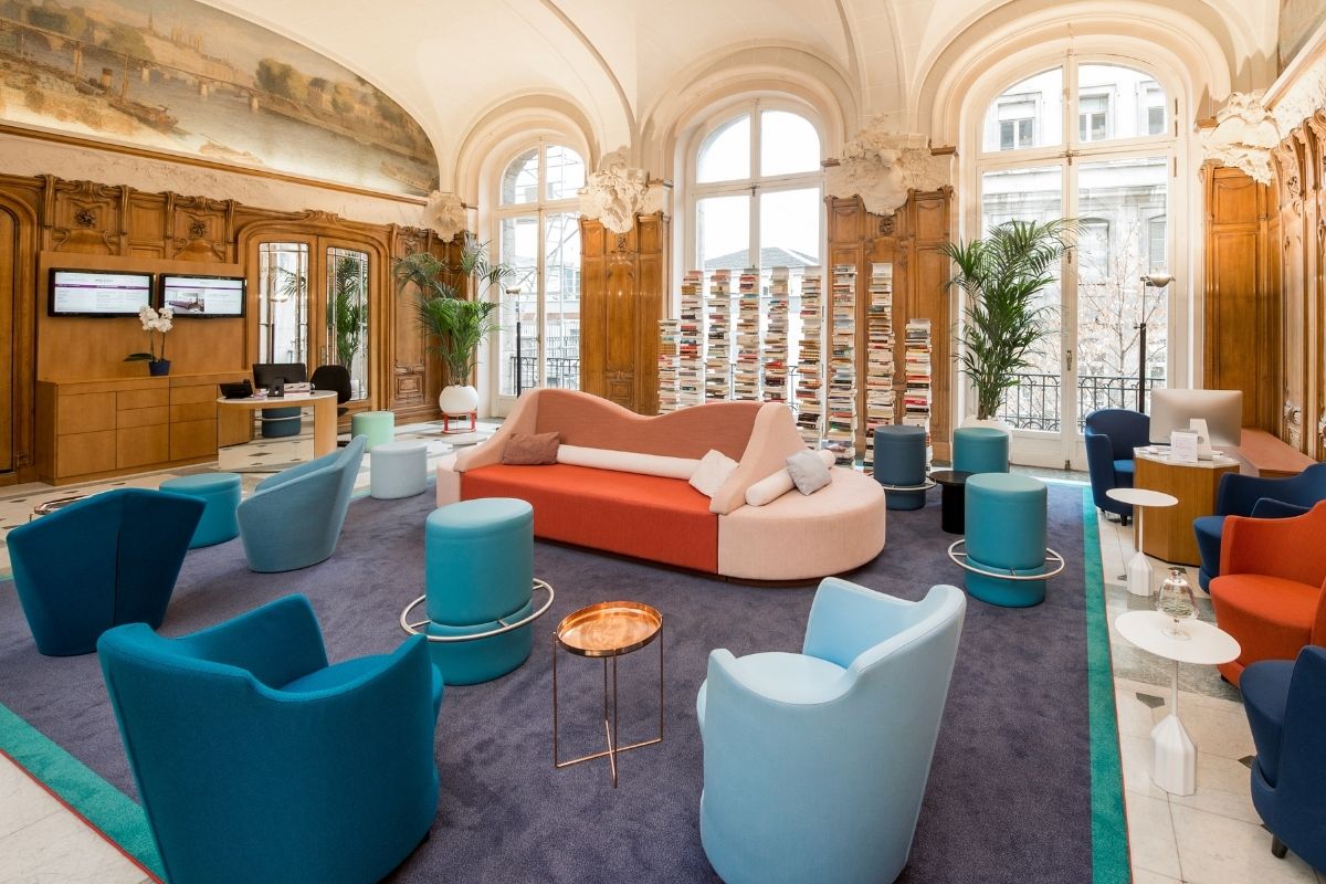 A wide angle view of a spacious hotel lobby lounge featuring vaulted ceilings, large arched windows, and wood-paneled walls, decorated with a central coral and pink curved sofa, colorful blue and teal armchairs, cylindrical ottomans, and tall stacks of bo