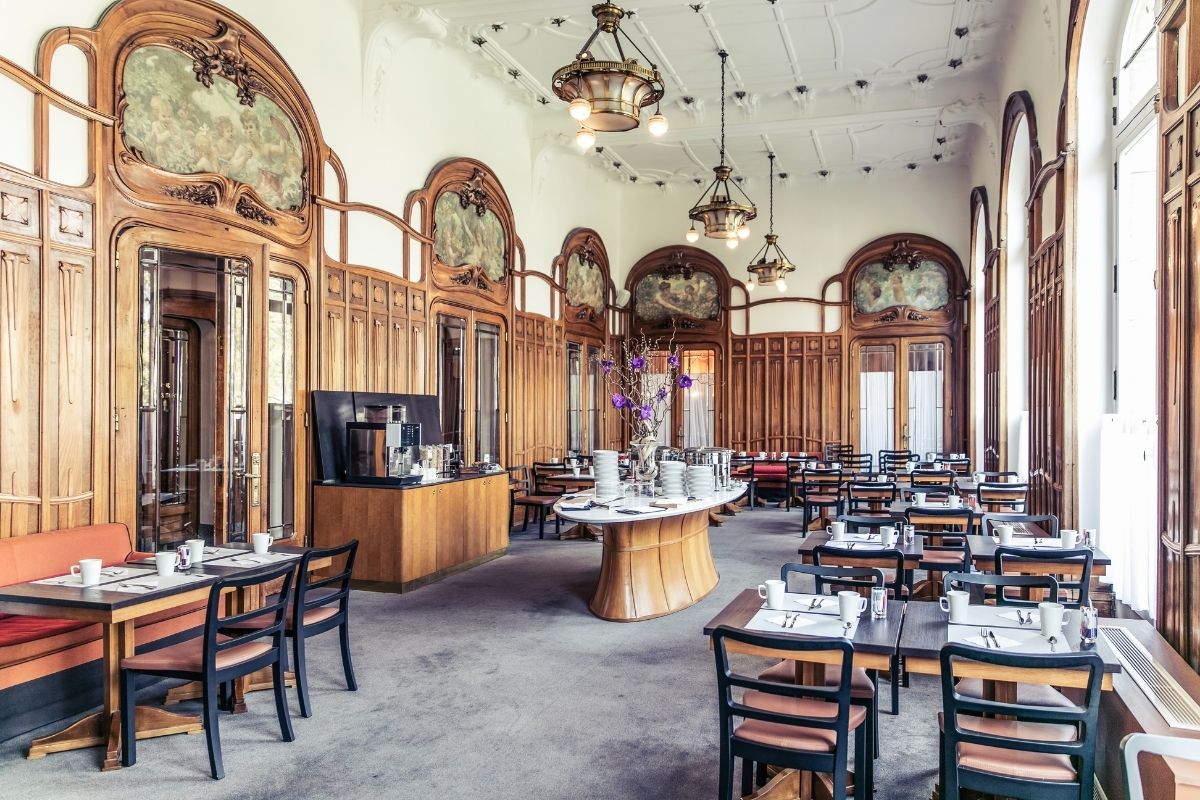 An alternate angle of the historic hotel dining hall highlighting ornate Art Nouveau wood paneling, classical artwork set above doorway arches, ornate ceiling details with brass chandeliers, and neat rows of set breakfast tables around a central buffet st