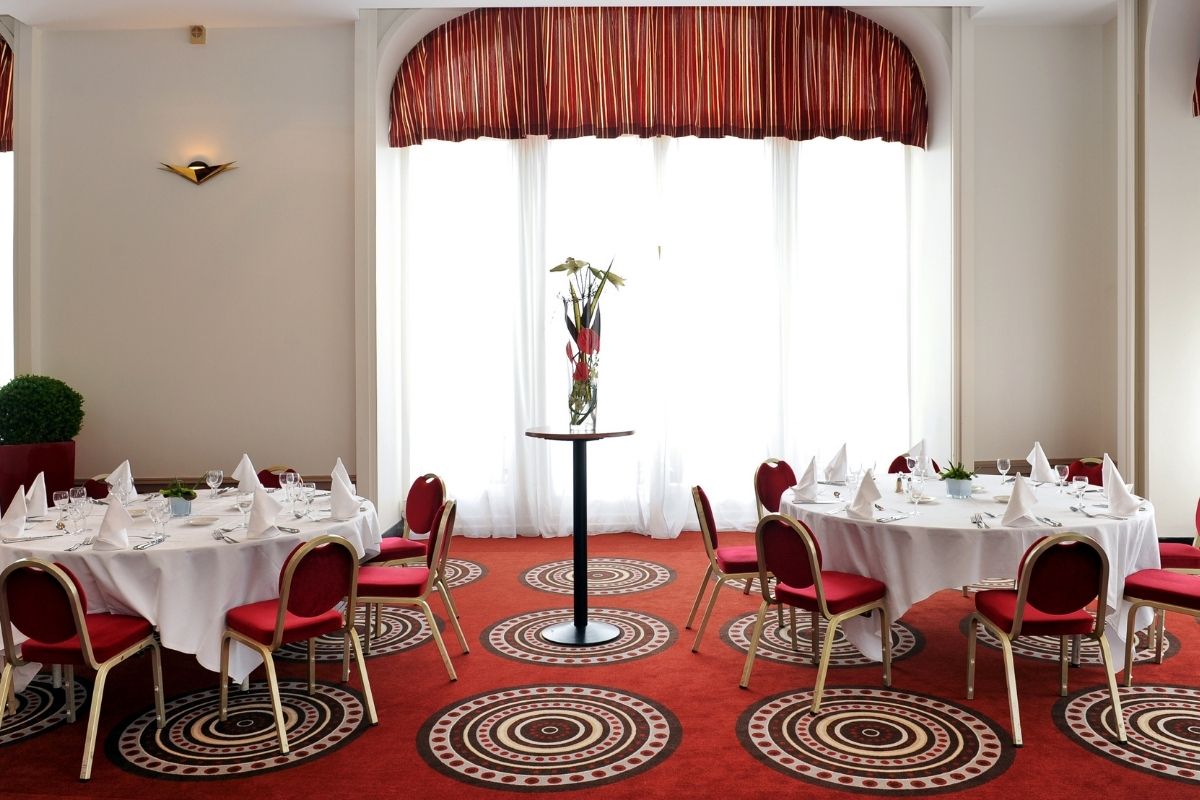 A banquet hall set with round, white-clothed dining tables, red upholstered chairs with gold frames, and a vibrant red carpet patterned with concentric circle motifs, illuminated by large sheer-curtained windows framed with striped red valances.
