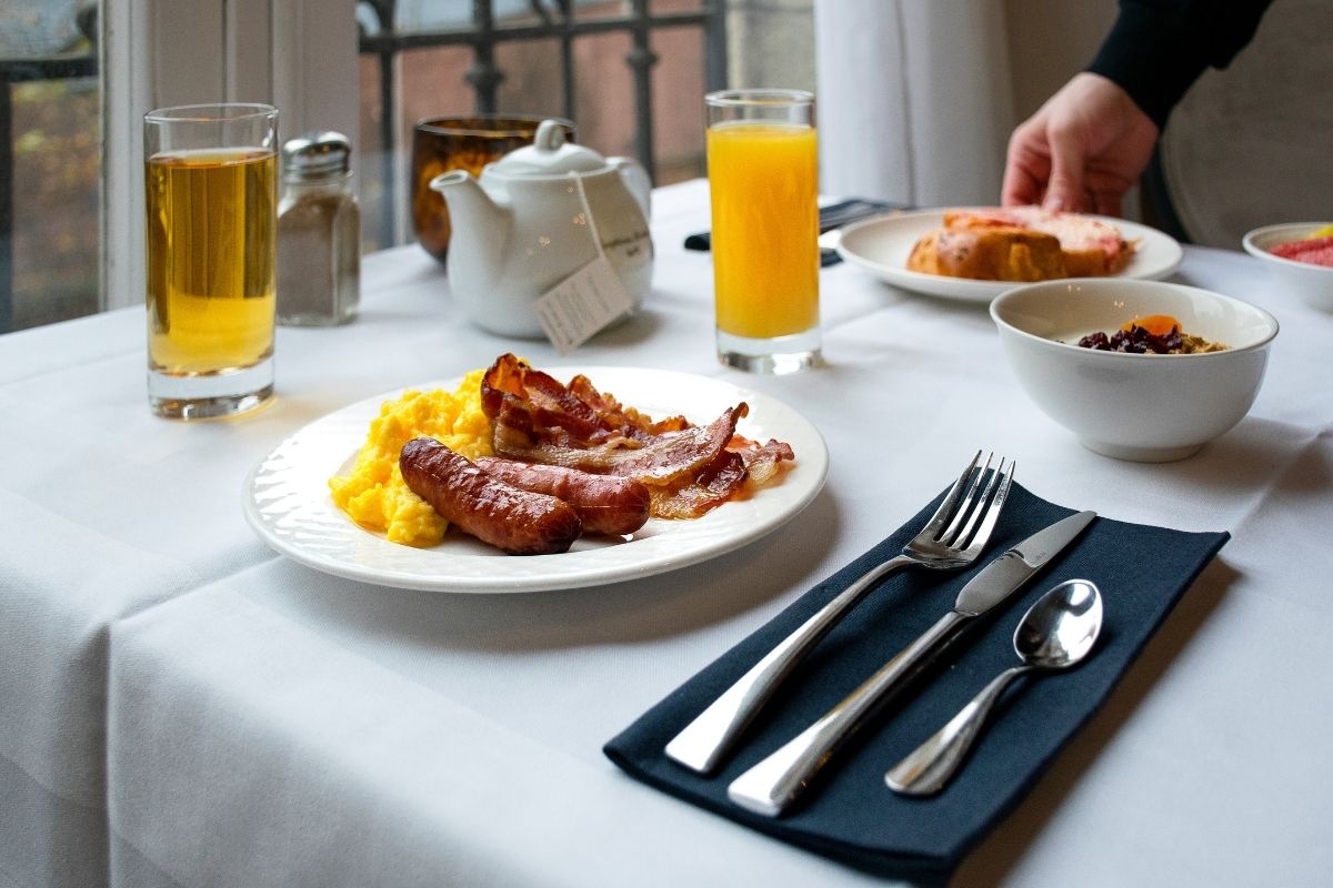 A white linen-draped hotel breakfast table set with a plate of scrambled eggs, crispy bacon, and sausages, alongside glasses of apple and orange juice, a white teapot, a bowl of oatmeal, and neatly arranged silverware on a dark napkin.