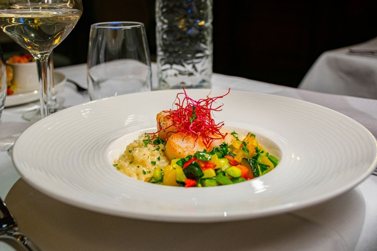 An elegantly plated gourmet dinner entry featuring seared scallops topped with vibrant red sprouts, served over creamy risotto and diced summer vegetables in a wide white bowl, accompanied by a glass of white wine.
