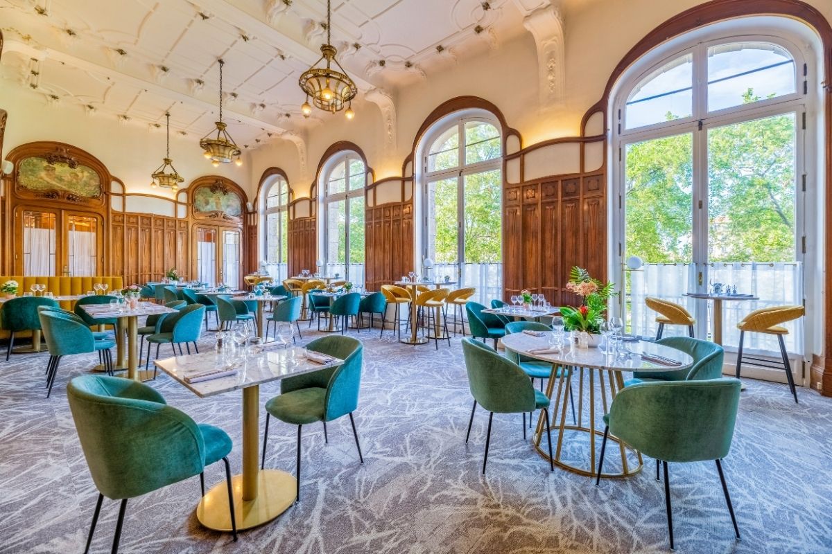 A bright hotel dining room with large arched windows overlooking greenery, combining historic Art Nouveau wood paneling and ornate ceiling moldings with modern teal velvet armchairs, marble-top tables, and mustard yellow accents.