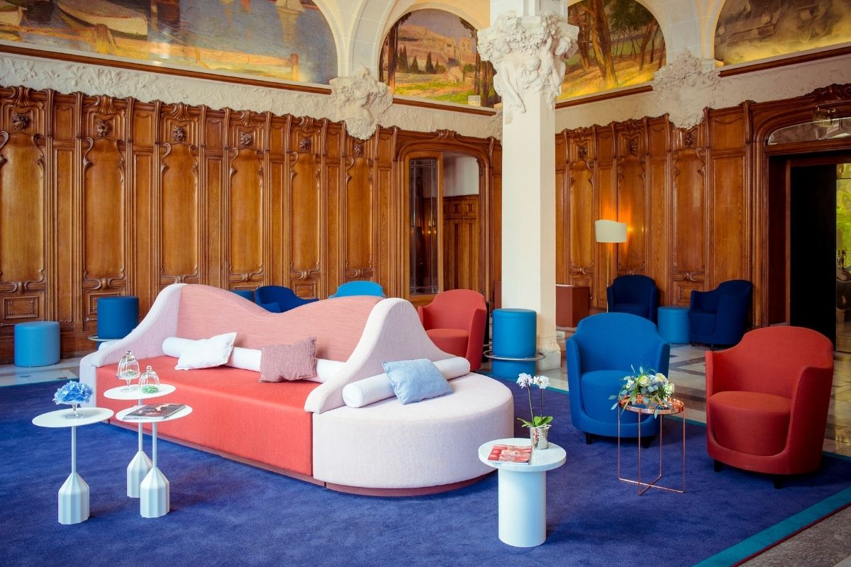 A grand hotel lobby lounge featuring classical carved wood wall paneling and ceiling murals juxtaposed with vibrant, modern furniture including a two-toned coral and blush curved sofa, bright blue and red armchairs, and small white side tables on a deep b