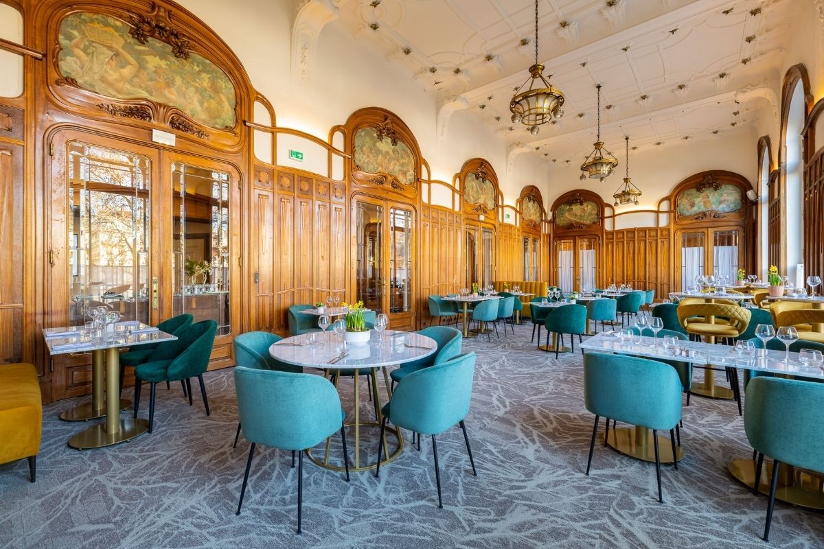A wide perspective of an elegant restaurant space featuring ornate Art Nouveau wood paneling, classic painted arched murals, intricate ceiling molding with hanging brass chandeliers, set with modern turquoise and teal velvet dining chairs around white mar