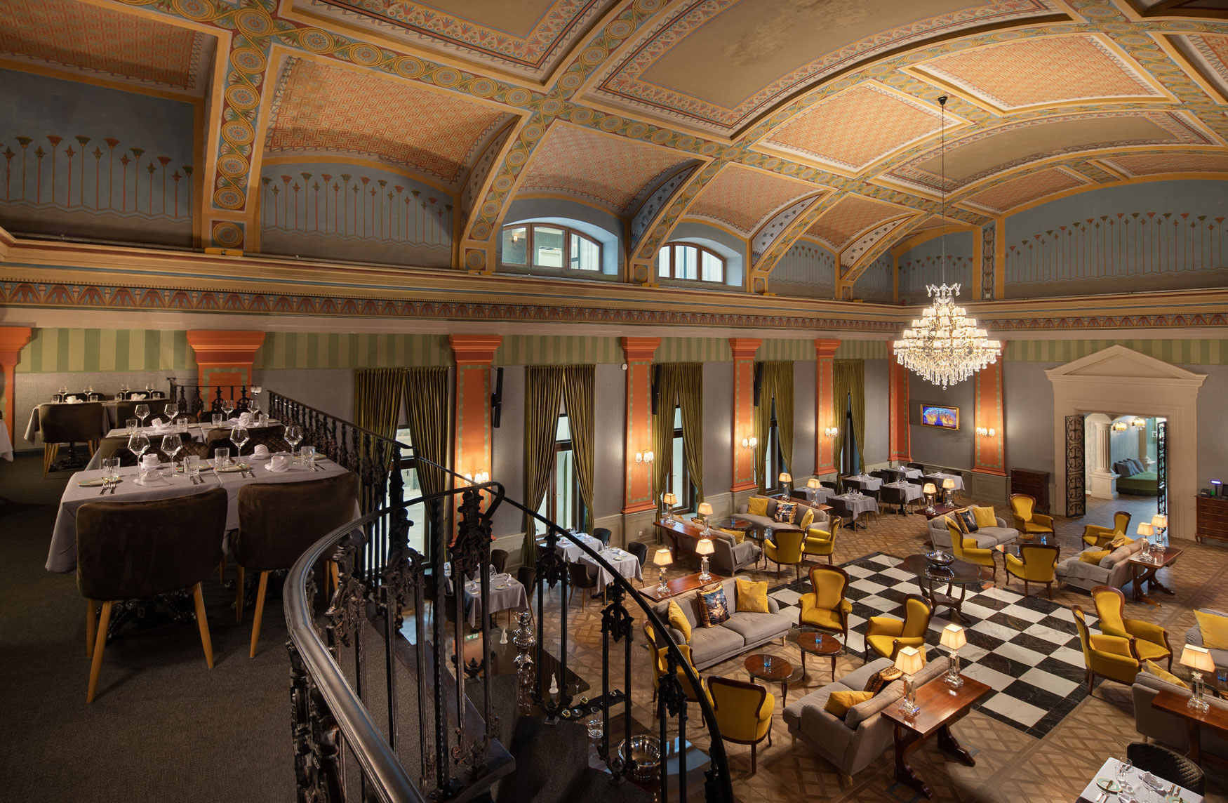 A high-angle view of a grand, opulent dining hall featuring a checkered floor, yellow armchairs, gray sofas, and a classic crystal chandelier beneath vaulted ceilings with intricate painted geometric patterns, viewed from an upper mezzanine dining area wi