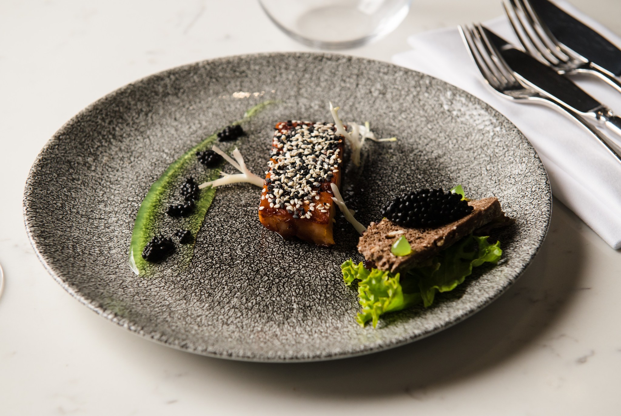 An elegantly plated fine dining appetizer featuring a sesame-crusted block of fish, dollops of black caviar served over a streak of green herb oil, and toasted rye crispbread topped with caviar, all presented on a textured gray ceramic plate.