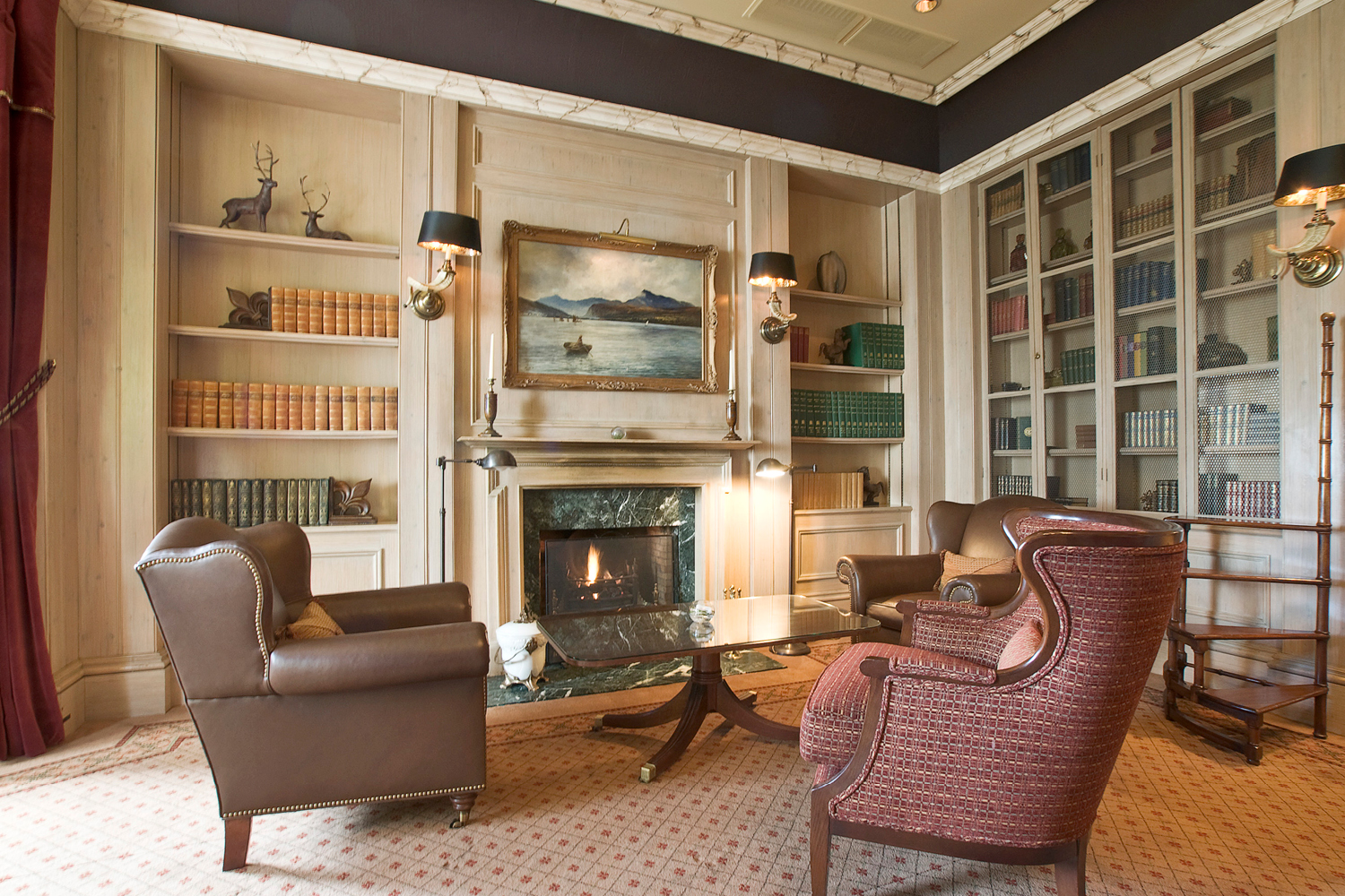 A cozy, sophisticated library sitting area centered around a marble-lined fireplace, featuring plush leather and patterned wingback armchairs, built-in wooden bookshelves, dark lampshades, and a classic landscape painting hung above the mantel.