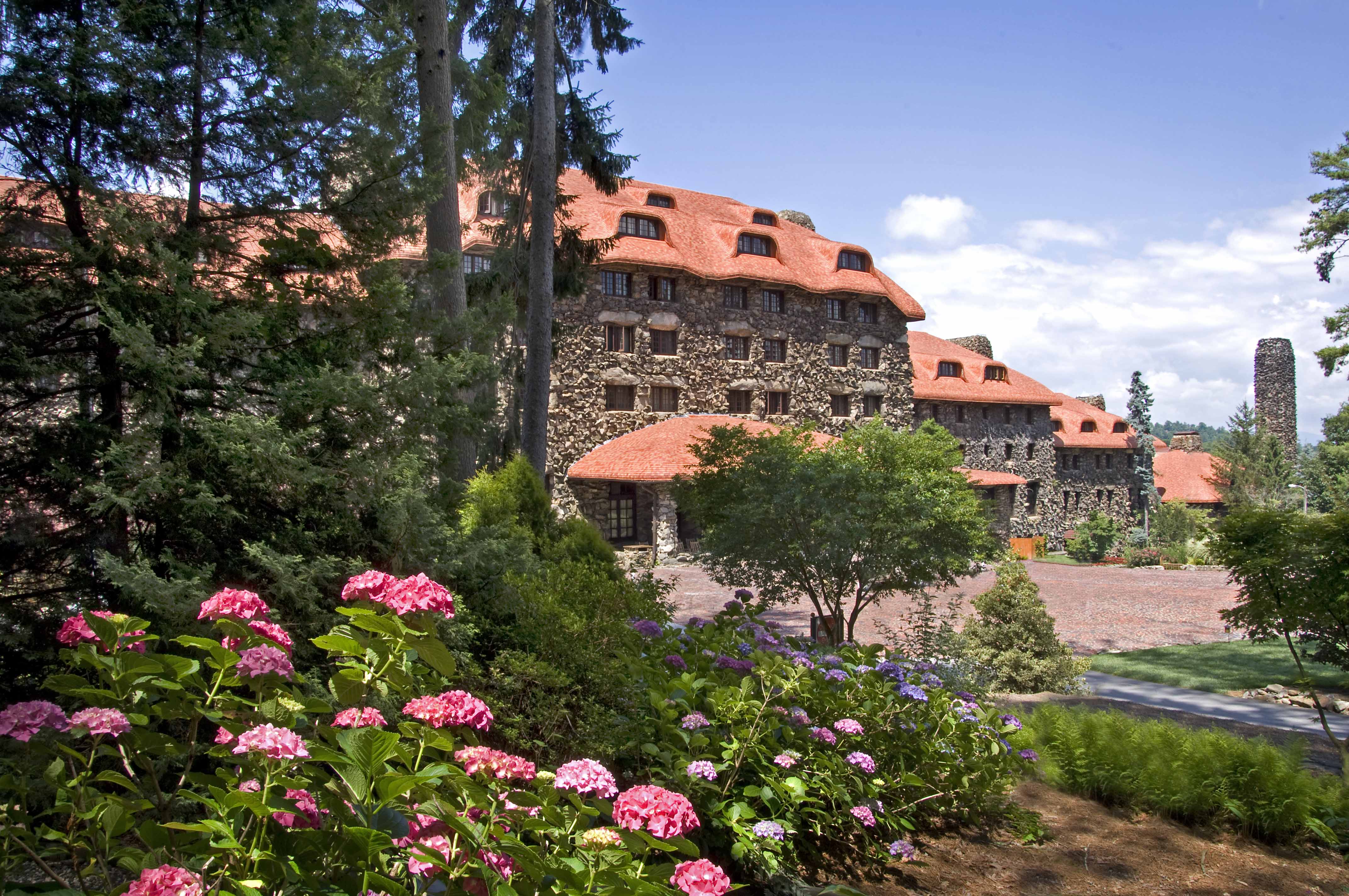 a large stone building with a red roof surrounded by trees and flowers