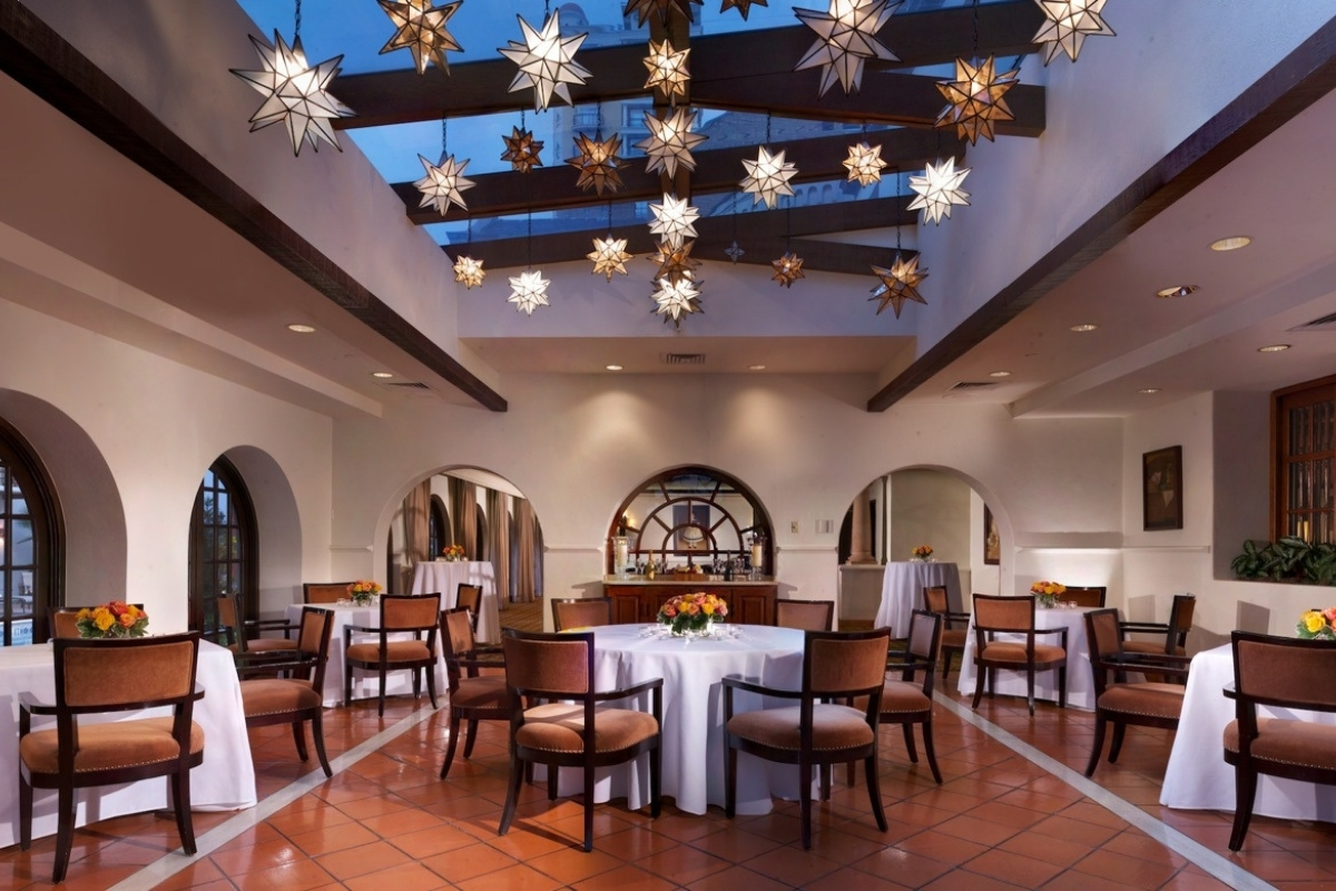 An elegant Spanish-style dining room featuring terracotta tile flooring, white arched doorways, dining tables with white tablecloths, brown velvet chairs, and hanging Morrocan star pendant lights from wooden ceiling beams.
