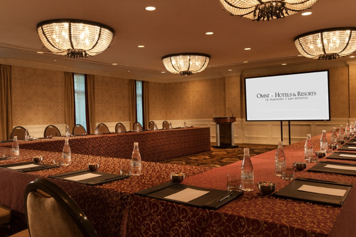 A formal meeting room set up with a U-shaped conference table, patterned tablecloths, crystal ceiling chandeliers, a podium, and a presentation screen displaying Omni Hotels & Resorts logo.