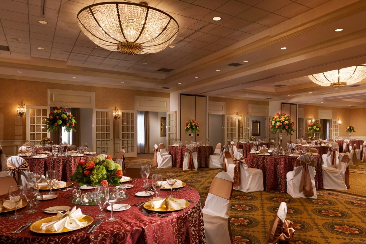 An elegant event ballroom set up for a banquet, featuring round tables with patterned red tablecloths, formal place settings with gold charger plates, vibrant floral centerpieces, white chair covers with gold bows, and flush-mount crystal chandeliers over