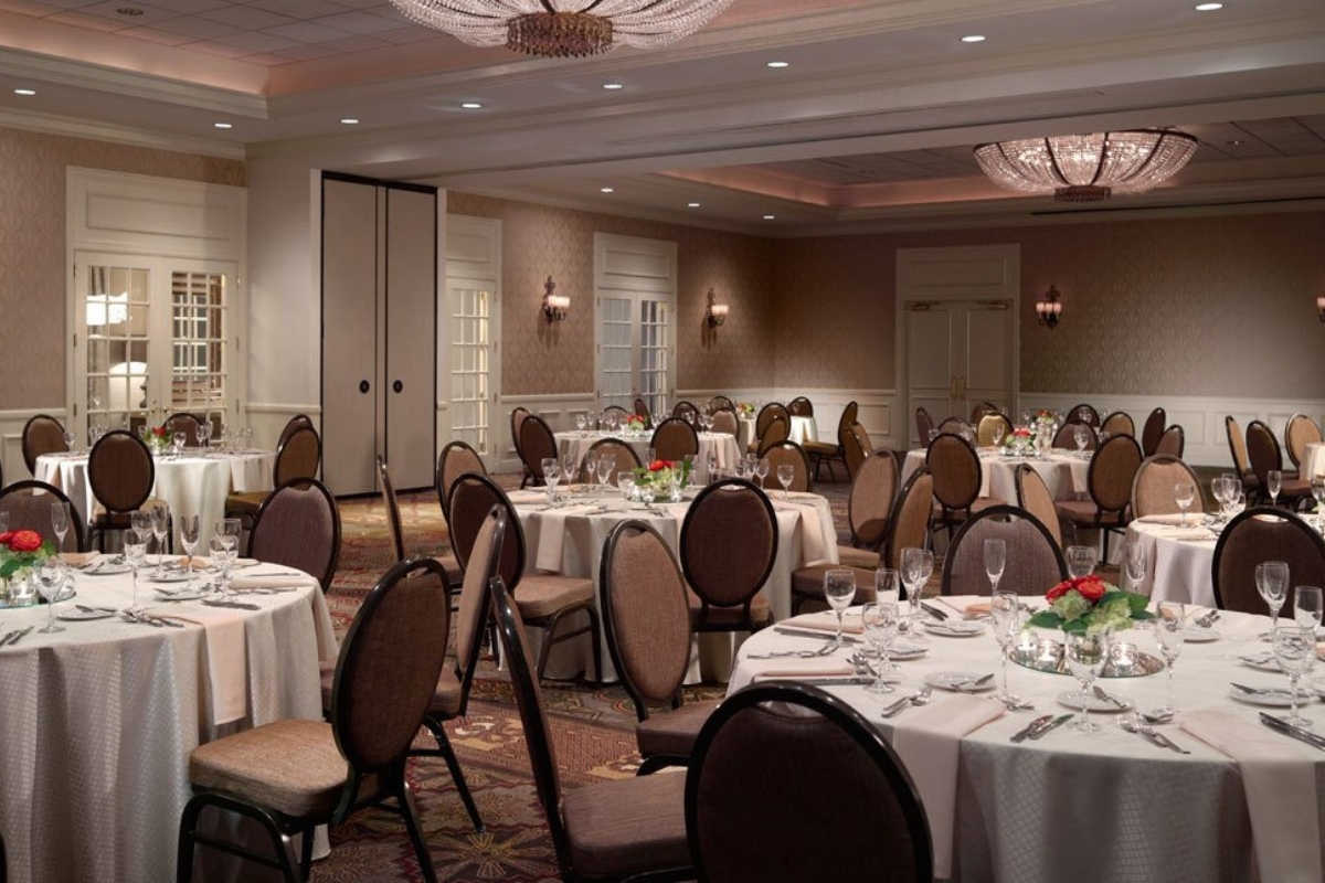 A hotel banquet hall set up with multiple round dining tables draped in cream-colored tablecloths, surrounding brown oval-back chairs, glassware, small floral centerpieces, and warm ceiling chandeliers.
