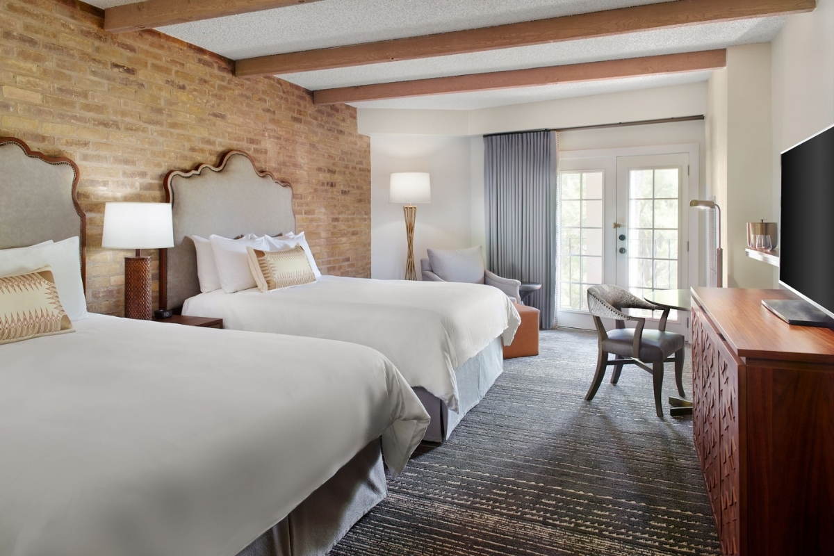 A hotel bedroom with an exposed brick wall, exposed wooden ceiling beams, two double beds with upholstered headboards, a TV console, an armchair, and French doors leading to a balcony.