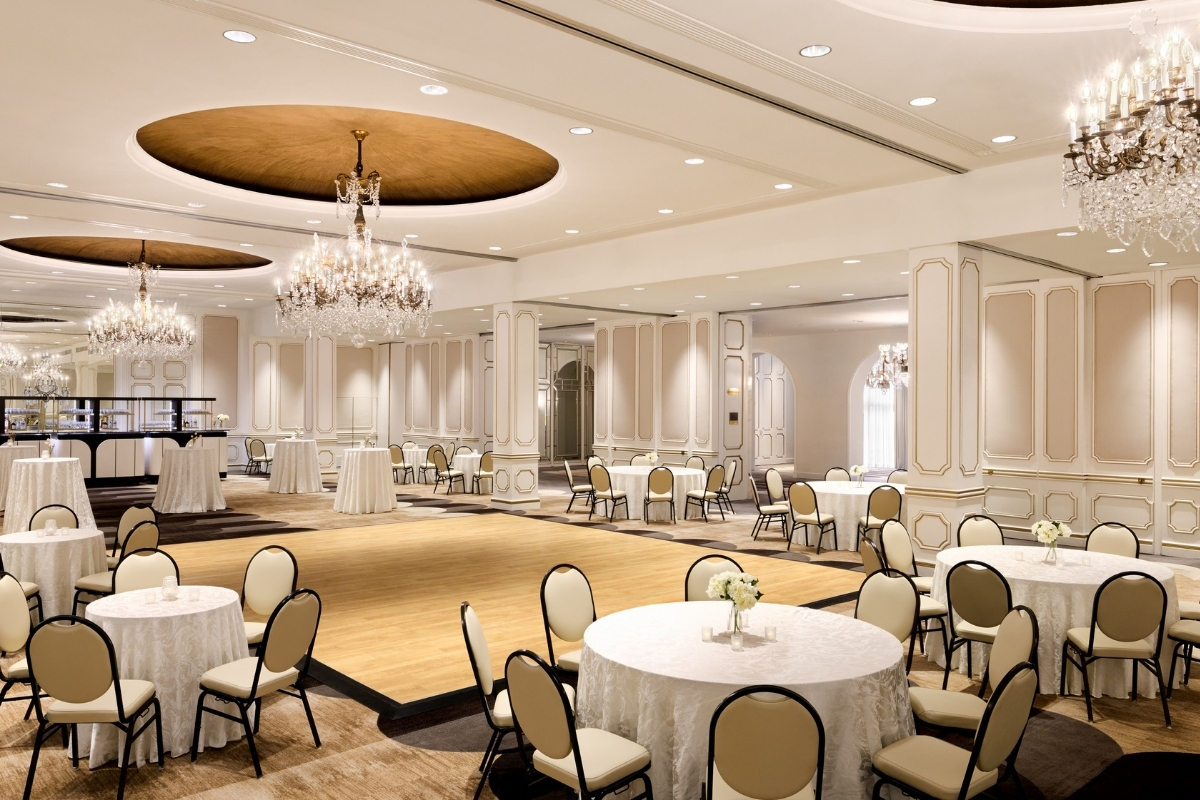 A brightly lit ballroom featuring ornate crystal chandeliers hanging from circular ceiling recesses, cream-colored paneled walls with gold trim, round dining tables, and a wooden dance floor in the center.