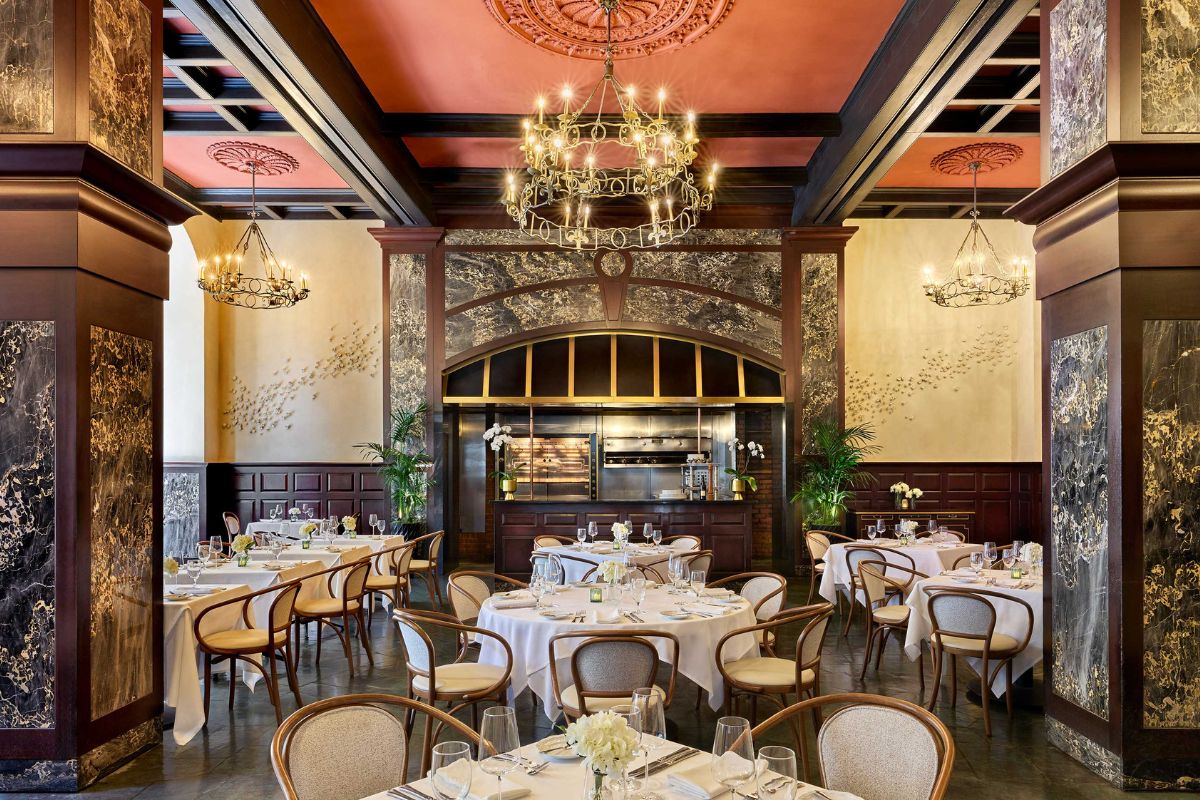 An upscale dining room featuring grand marble pillars, dark wood paneled walls, a peach-toned ceiling with brass chandeliers, and round white-linen tables with bentwood chairs surrounding an open kitchen area.