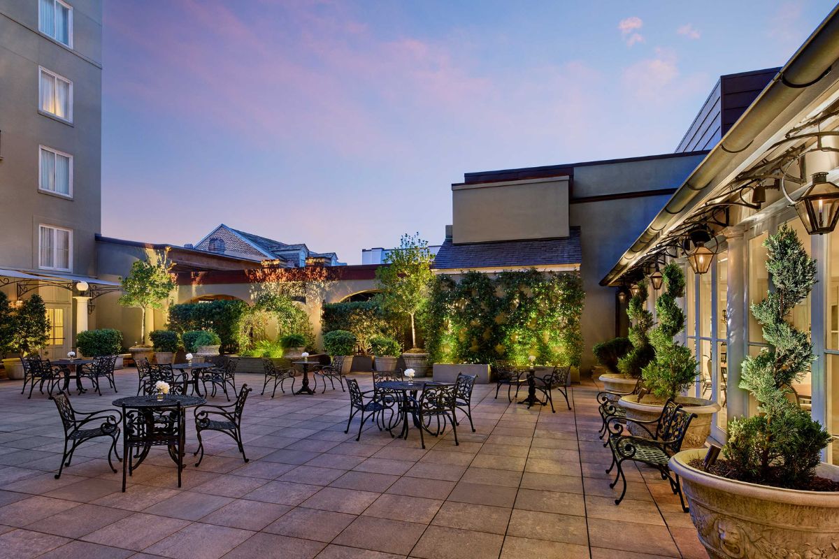 A spacious open-air hotel patio at dusk, featuring stone paving, wrought-iron dining tables and chairs, lush potted topiary trees, green ivy-covered walls, and warm hanging lanterns.