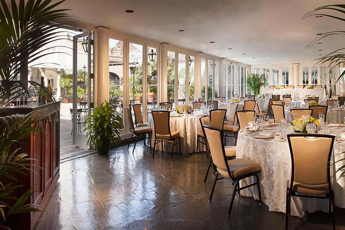 A bright sunroom dining hall with floor-to-ceiling glass doors opening to an outdoor courtyard, filled with round tables set with white patterned tablecloths and elegant high-back dining chairs.