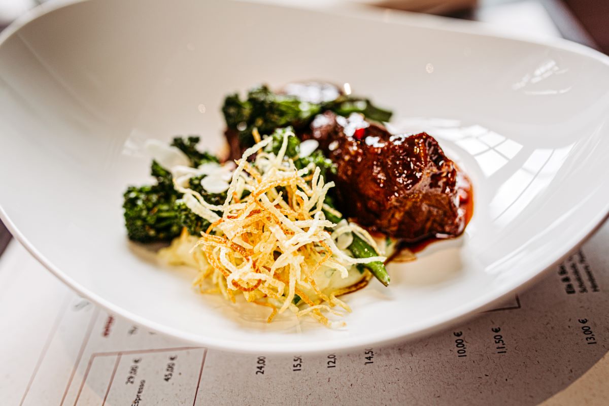 A gourmet plated dish served in an asymmetrical white bowl, featuring tender glazed braised beef, charred broccolini, creamy mashed potatoes, and a nest of crispy shoestring potato straw.