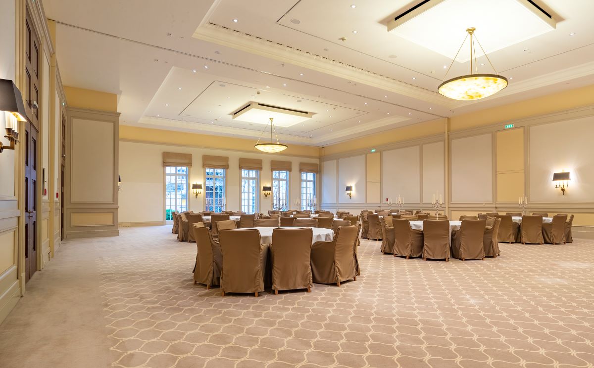 A spacious, elegant banquet hall set up for an event, featuring round dining tables with white tablecloths and beige chair covers, wide patterned carpeting, high ceilings with modern pendant chandeliers, and large French windows along the far wall.