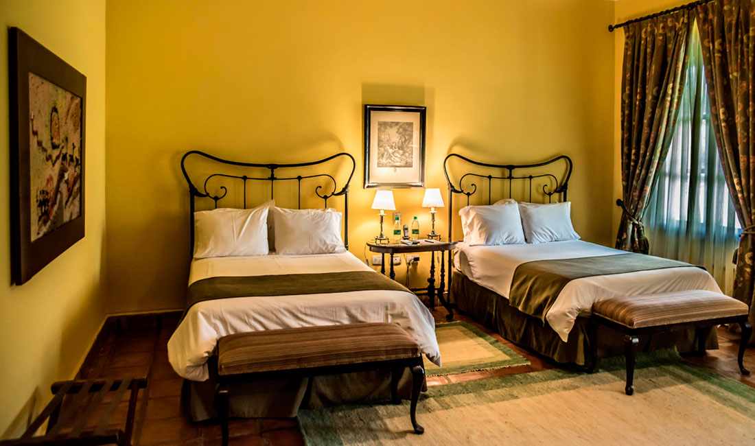 A warm hotel bedroom with yellow walls and tile flooring, featuring two beds with wrought-iron headboards, dark green throw blankets, upholstered benches at the feet of the beds, and a shared nightstand with small lamps.
