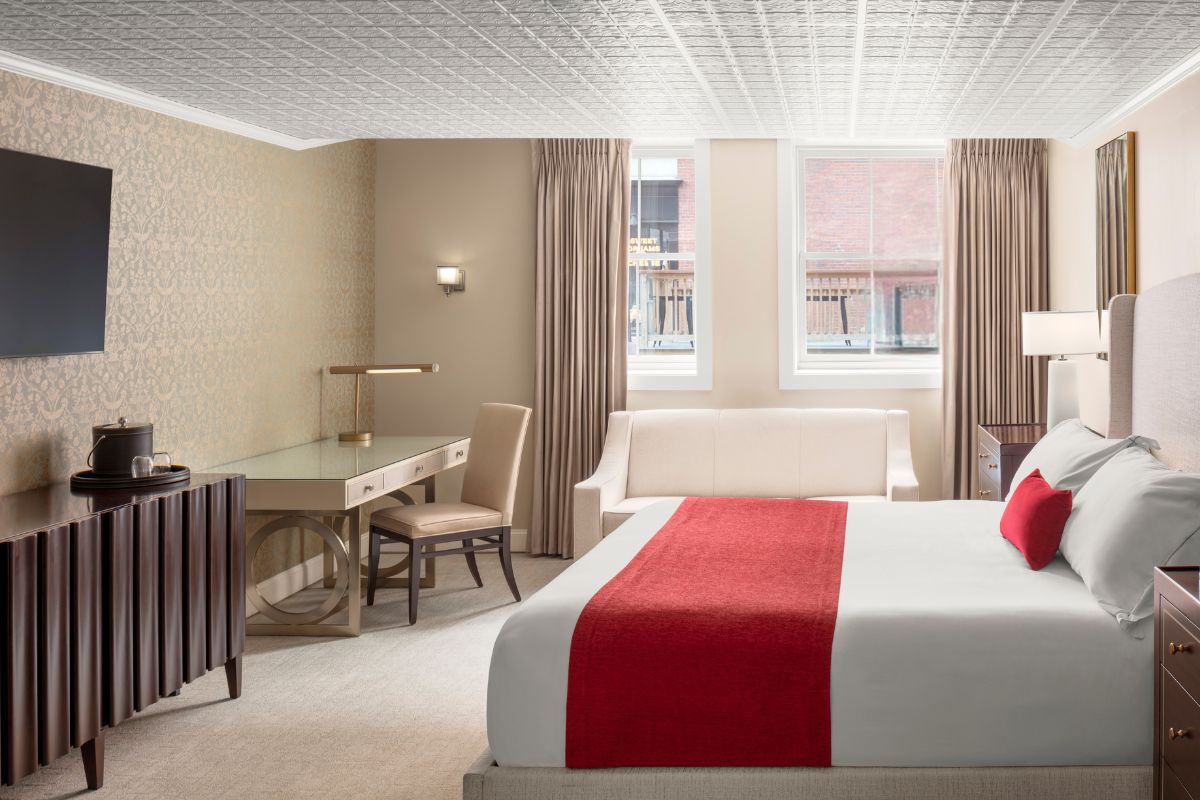 Modern hotel room featuring a king-size bed with white linens and a red accent runner, a glass-top work desk with a chair, a white sofa by two sash windows, and a dark wood credenza under a wall-mounted TV.