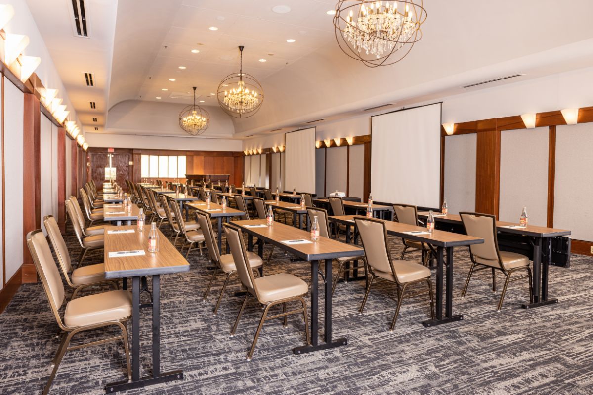 Large, organized conference room set up in classroom style with long wood-topped tables, padded beige chairs, hanging crystal orb chandeliers, and projection screens along the wall.