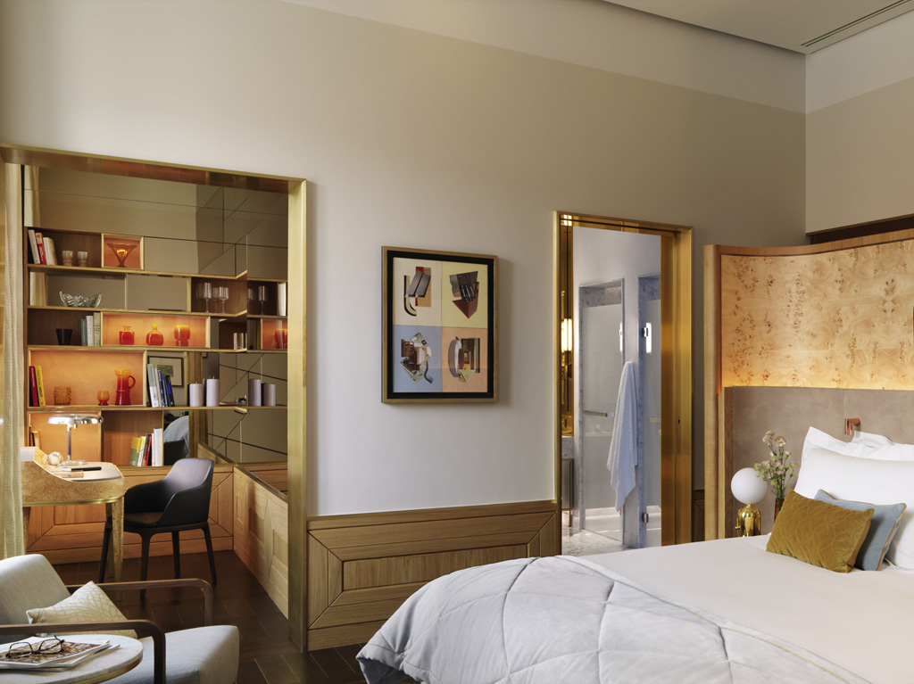 A luxurious hotel suite interior showing a bed with a warm wood headboard on the right, an adjoining study alcove with a desk and mirrored shelves on the left, and an open door leading into a sleek bathroom.