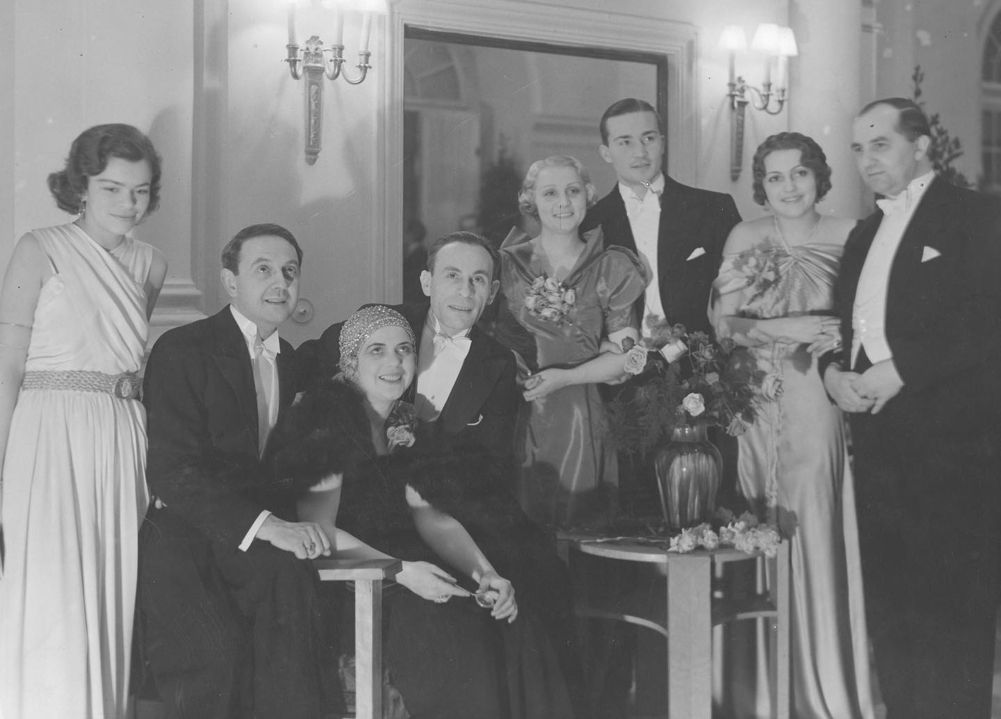 A vintage black-and-white group portrait of elegantly dressed men in tuxedos with bow ties and women in formal evening gowns and 1930s style attire gathered indoors around a small side table.