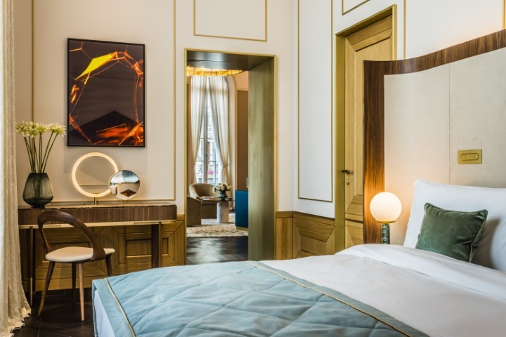 A stylish hotel bedroom with a bed featuring a sage green accent pillow in the foreground, a vanity desk with an illuminated ring mirror on the left, and an doorway leading to an adjoining living area.