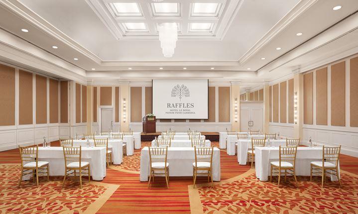Large, elegant ballroom set up classroom-style with white-clothed tables and gold chiavari chairs, facing a stage with a podium and a projection screen displaying 'RAFFLES HOTEL LE ROYAL PHNOM PENH CAMBODIA'.
