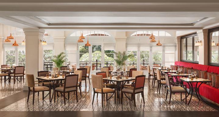 Brightly lit hotel dining room with patterned tile flooring, red plush banquette seating along one wall, wooden dining sets, copper pendant lamps, and large windows looking out to greenery.