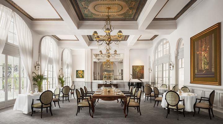 Grand fine-dining restaurant interior with high coffered ceilings, ornate chandeliers, hand-painted ceiling panels, tall arched windows with sheer drapes, a long wooden central dining table surrounded by smaller tables, and a black grand piano in the back