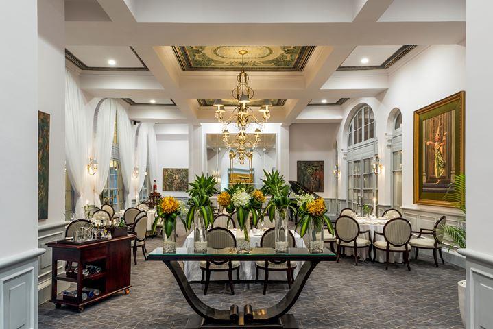 Elegantly decorated hotel dining room featuring a foreground dark console table with vibrant floral arrangements, grey carpet, round-backed dining chairs set around white-clothed tables, an ornate chandelier, coffered ceiling, and a large framed artwork o