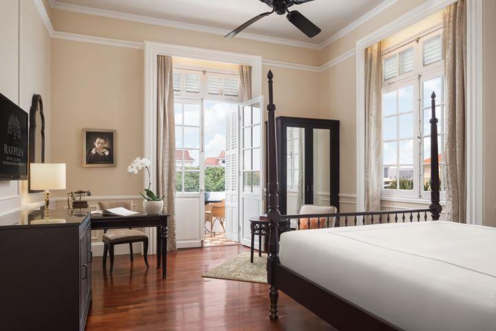 Colonial-style hotel suite featuring a four-poster bed, dark wood furniture, polished hardwood floors, french doors opening to a balcony, and warm cream walls.