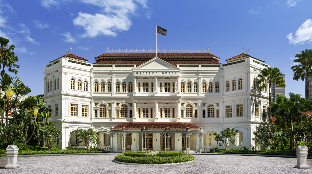 a large white building with a flag on top with Raffles Hotel in the background, beautiful blue sky.