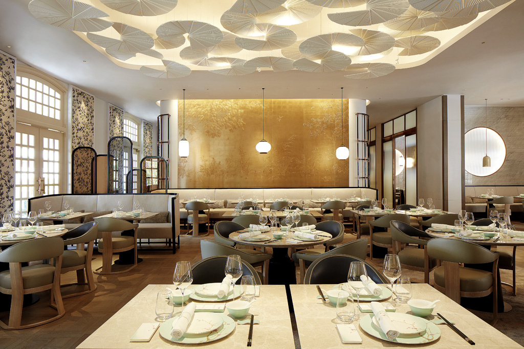 A modern, bright luxury Asian dining room featuring light wood tables set with delicate tableware, modern curved armchairs, a gold feature wall with pendant lights, and a white fan-shaped ceiling installation.