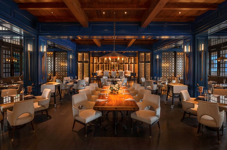 A warm, atmospheric dining room with rich royal blue paneled walls, exposed wood ceiling beams, cream-colored keyhole armchairs surrounding a central live-edge wooden table, and illuminated wine displays in the background.