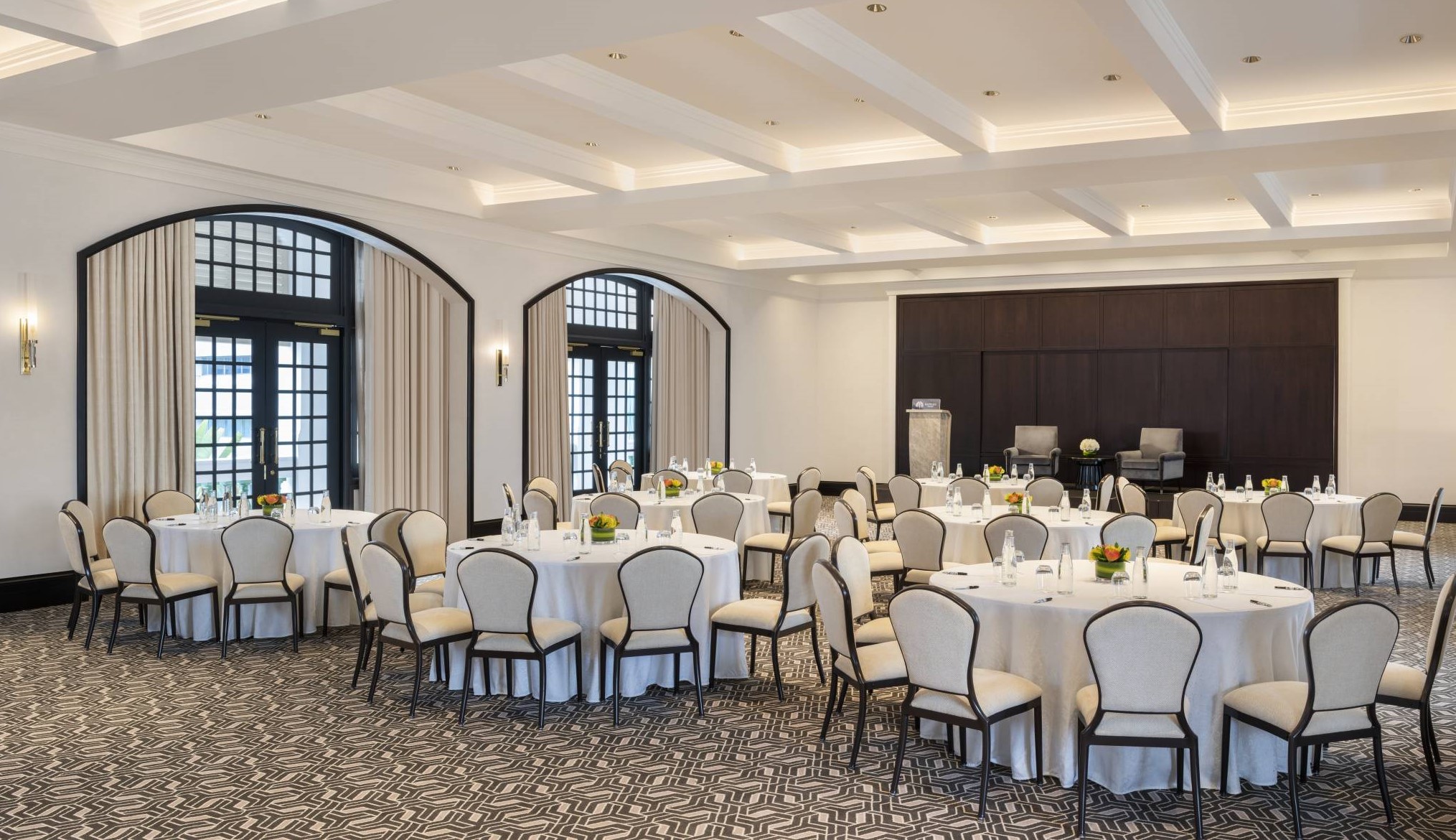 A bright, spacious event room set with round tables draped in white linens and surrounded by cream upholstered chairs, featuring coffered ceilings, patterned carpet, arched glass doors, and a dark wood accent wall with a podium.