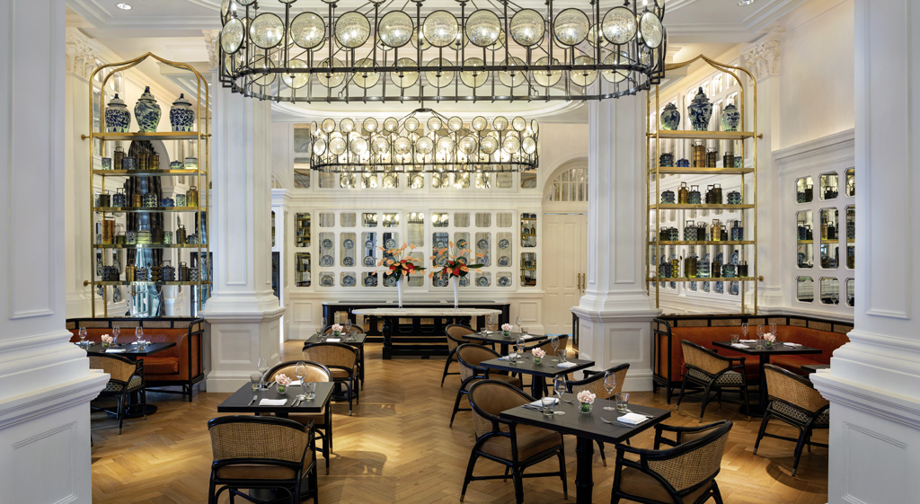 A stylish dining space with light wood herringbone floors, dark dining tables, and woven cane chairs, featuring brass shelving units filled with blue-and-white porcelain and large oval glass disc chandeliers overhead.