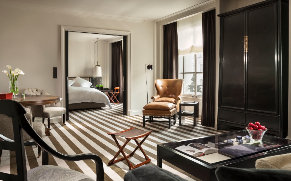Spacious luxury hotel suite with bold striped carpeting, a living area with a leather armchair, black armoire, and coffee table, leading into an adjoining bedroom.