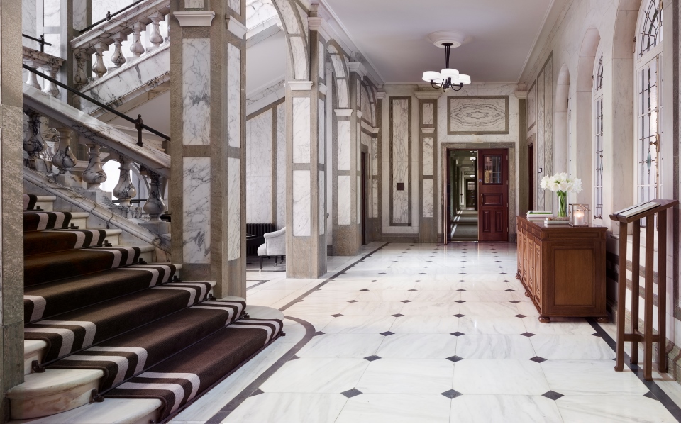 Grand hotel lobby featuring marble walls and columns, a grand marble staircase with a striped runner, polished marble floor tiles with dark diamond accents, and a wooden console table.