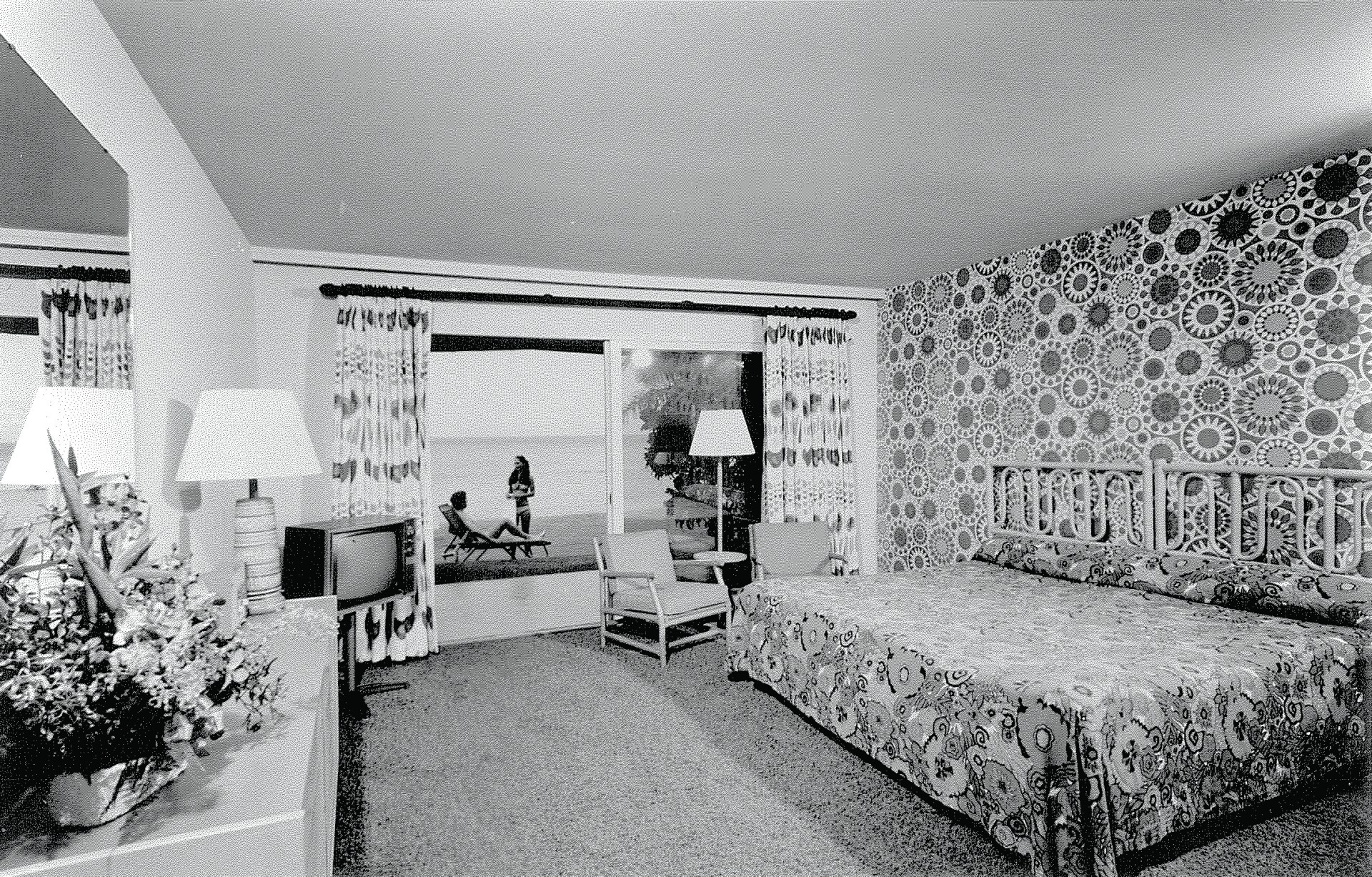 A historical black-and-white interior photograph of a 1960s-era hotel room featuring bold geometric wallpaper, a bed with a bamboo headboard and patterned bedspread, a small television, and sliding glass doors revealing guests on a oceanfront terrace.