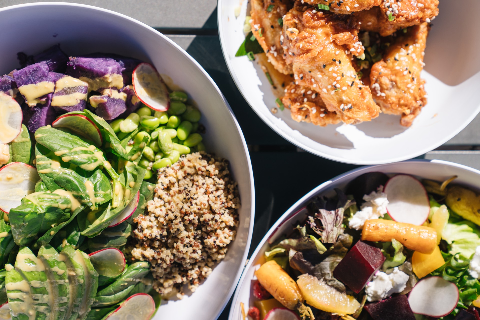 A top-down view of three white serving bowls containing a quinoa and avocado grain bowl with purple sweet potatoes, sesame-glazed fried chicken wings, and a roasted beet salad with goat cheese.